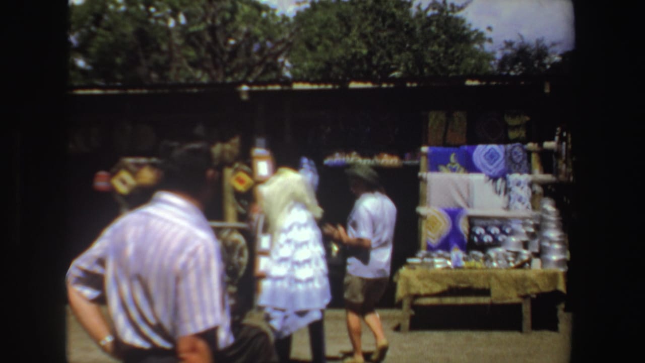1969 South Africa home-movie frame showing visitors near outdoor craft tables with patterned fabrics, shelves, and hanging goods.