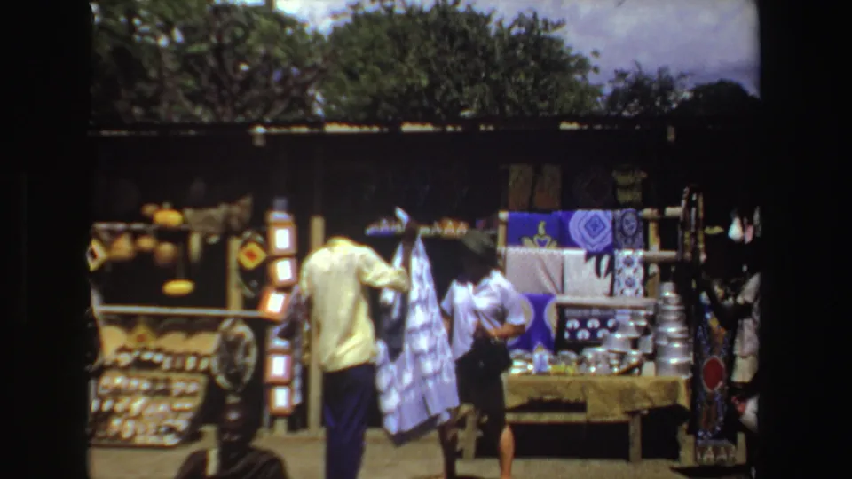 Home-movie footage from South Africa in 1969 shows visitors browsing a bright outdoor market stall with textiles, shelves, and craft goods.