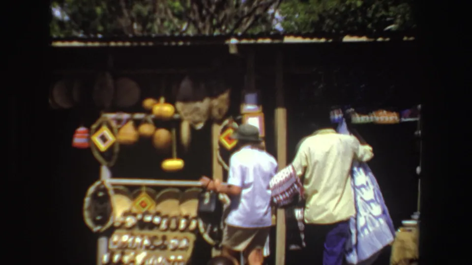 A late-1960s South Africa market frame shows visitors from behind near woven or wooden items, hanging objects, and patterned textiles.