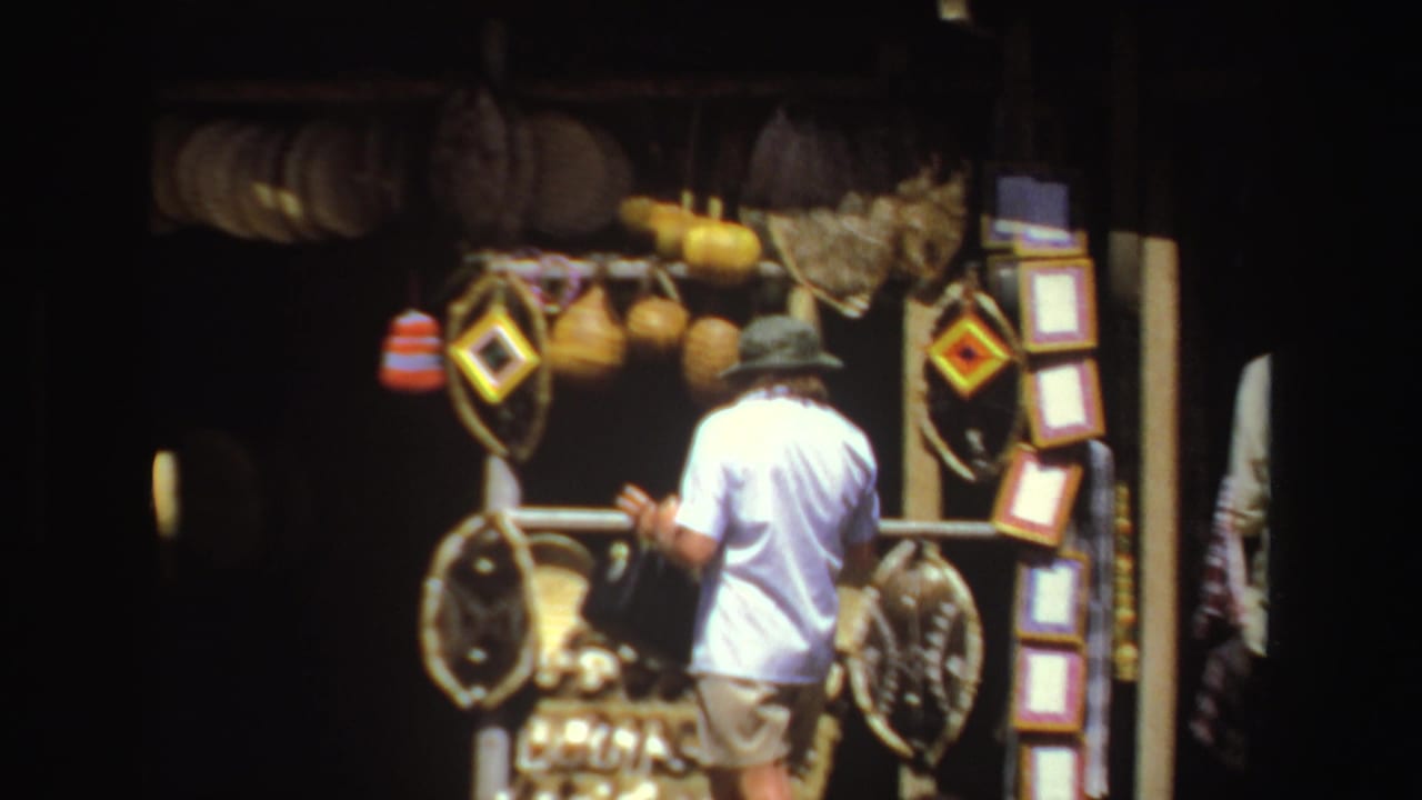 1969 South Africa home-movie frame of a person browsing hanging baskets, gourds and craft pieces at a shaded market stall.