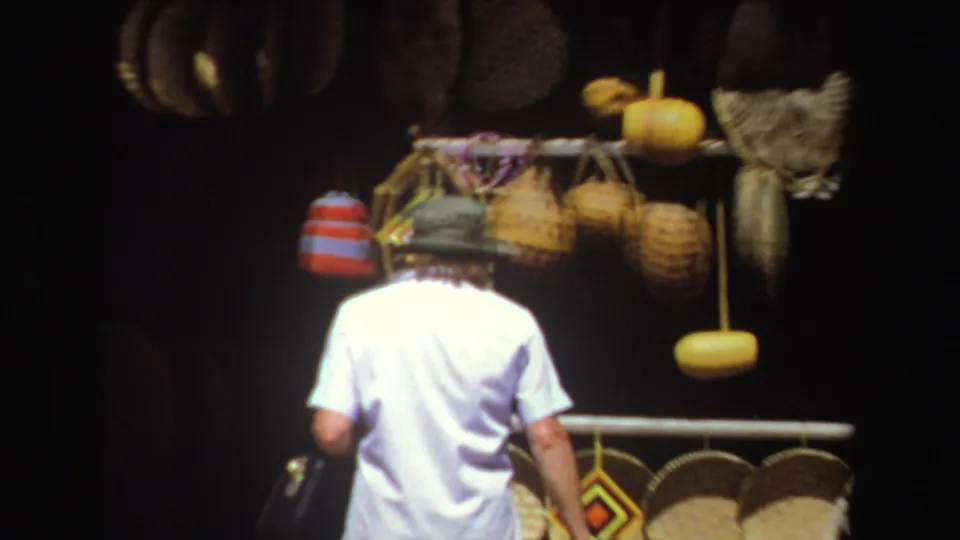 In this 1969 South Africa frame, a visitor in a white shirt stands under hanging baskets and gourds at a craft stall.