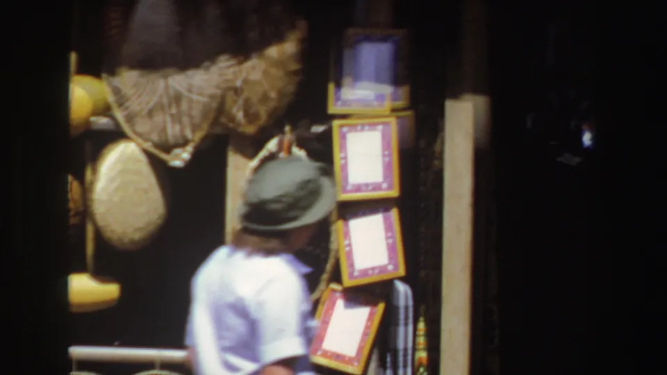 1969 South Africa home-movie frame with a visitor beside hanging woven goods and a vertical stack of framed craft pieces.