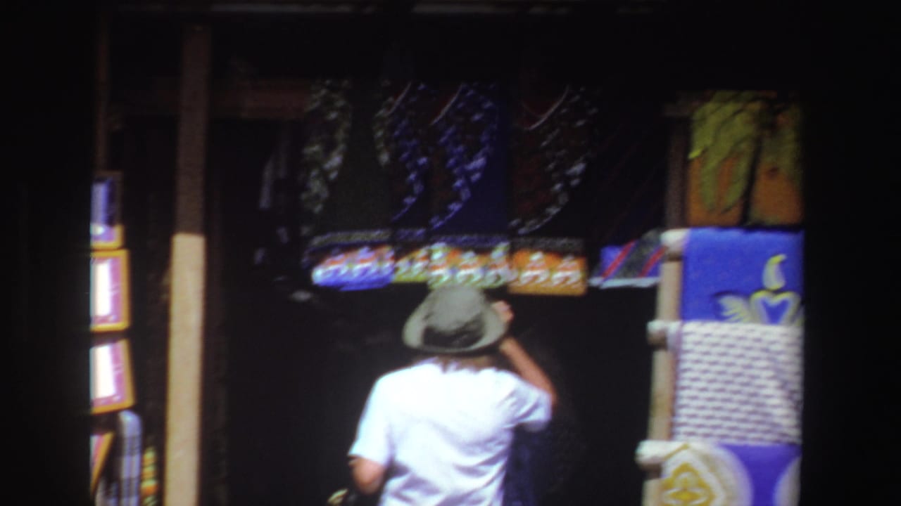 Person in a green hat walks past textiles and framed items at a market stall.
