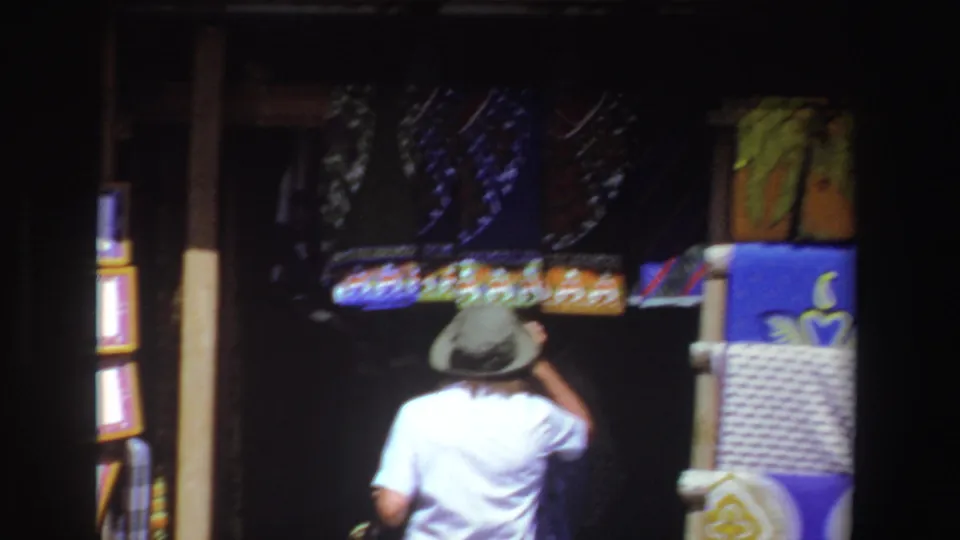 The shopper is seen from behind near colorful textiles, framed pieces, and woven goods in 1969 South Africa market footage.