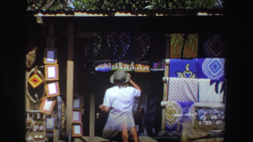 A 1969 South Africa outdoor market frame shows a rear view of a shopper examining colorful textiles and craft displays.