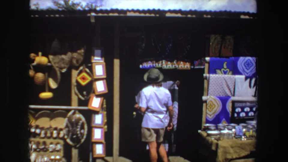 The final frame shows the shopper from behind beside bright textiles and a table of small market goods in South Africa, 1969.