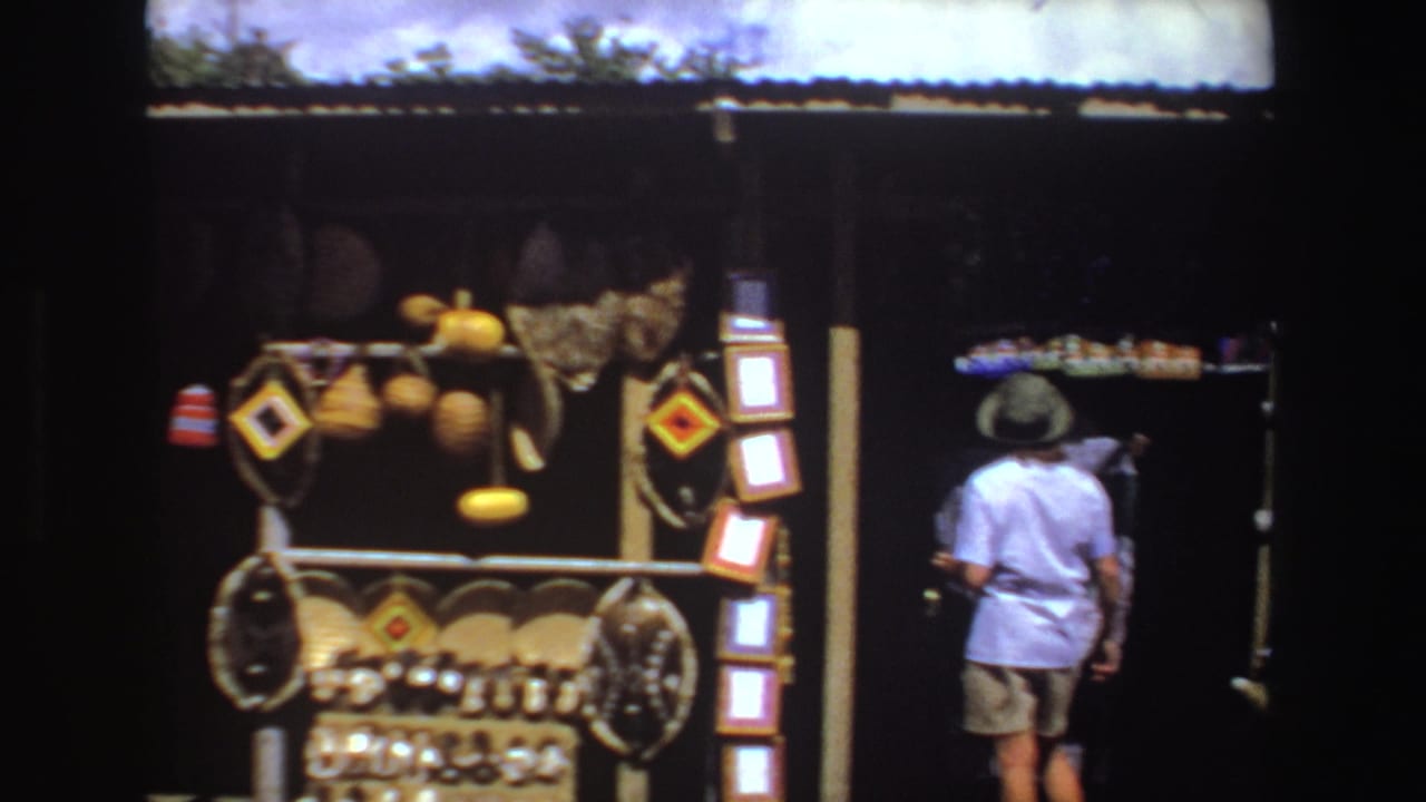 1969 South Africa 8mm frame showing a roadside stall with woven baskets, gourds, patterned textiles, and a passerby in a white shirt and hat.