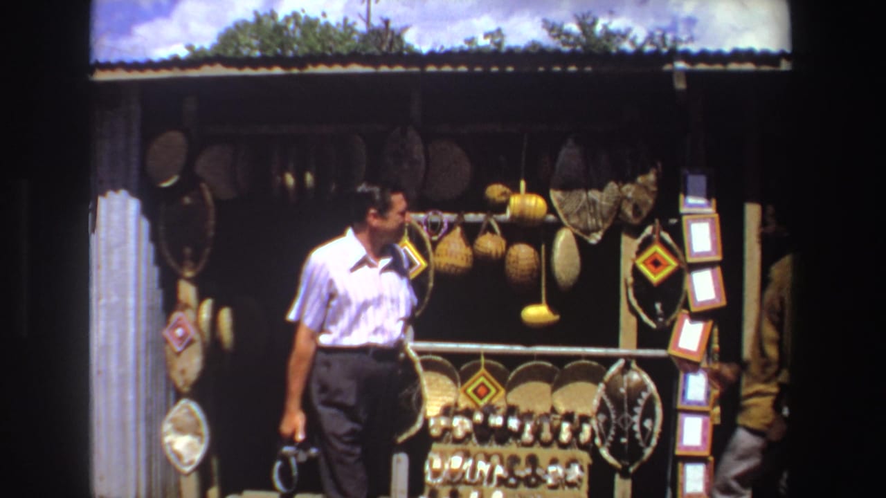 Man in a white striped shirt stands near a roadside stall filled with baskets and hanging goods.