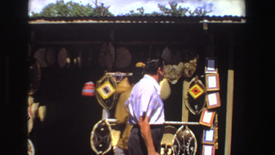 1969 South Africa 8mm frame showing a man browsing at a roadside craft stall while another person stands partly hidden near the display.