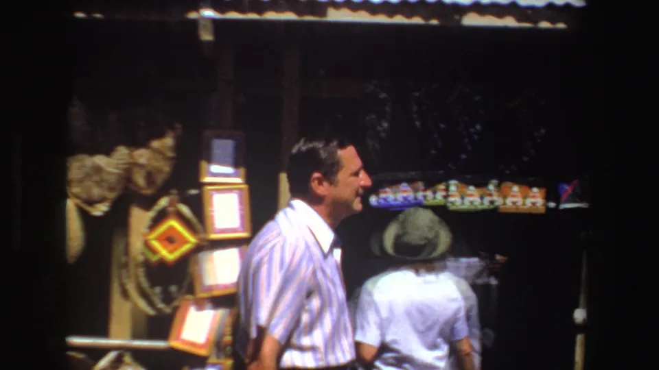 1969 South Africa 8mm frame with a smiling man in a white striped shirt and a hat-wearing visitor near a stall of baskets and textiles.