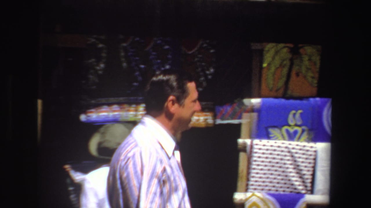 In 1969 South Africa footage, a man in a striped button-down looks toward bright patterned scarves at an outdoor market stall.