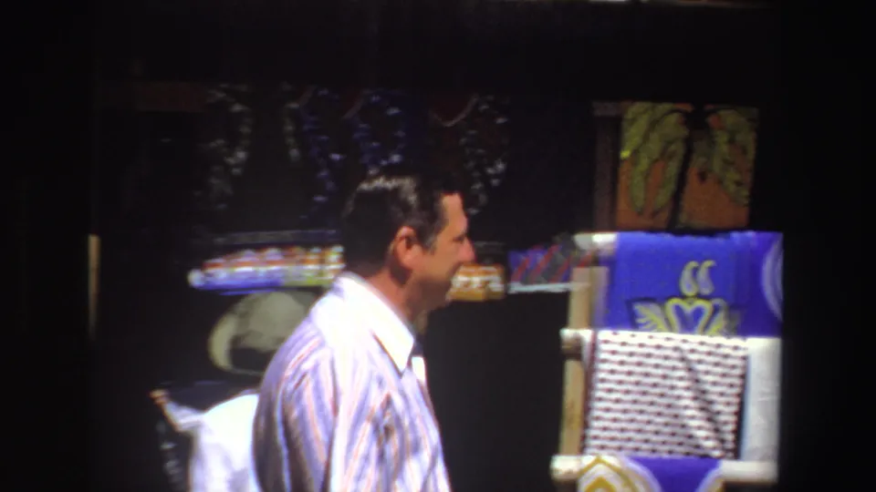 In 1969 South Africa footage, a man in a striped button-down looks toward bright patterned scarves at an outdoor market stall.