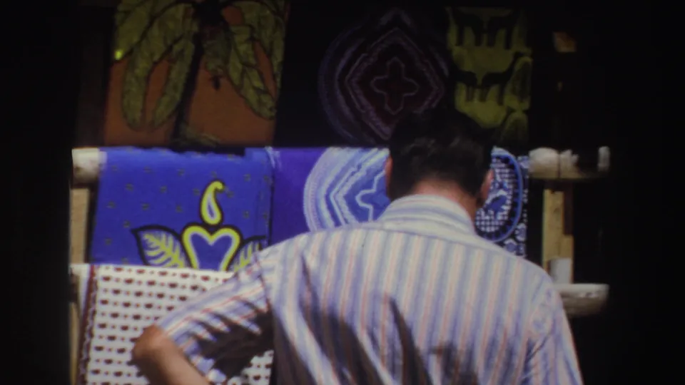 A closer rear view shows the striped-shirted man pausing before layered blue, white, and yellow textiles in the 1969 market footage.