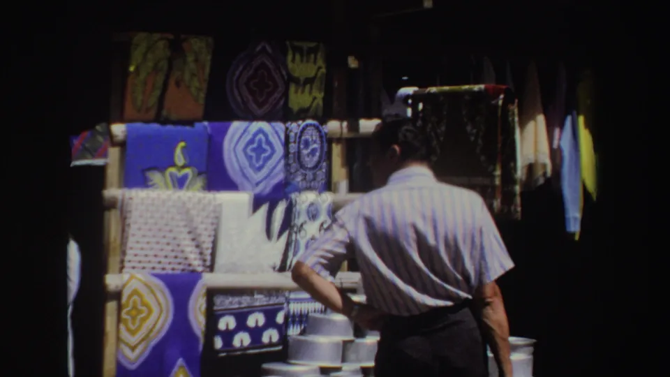 The man stands near the textile rack with one hand on his hip, surrounded by hanging fabric displays in outdoor market shade.