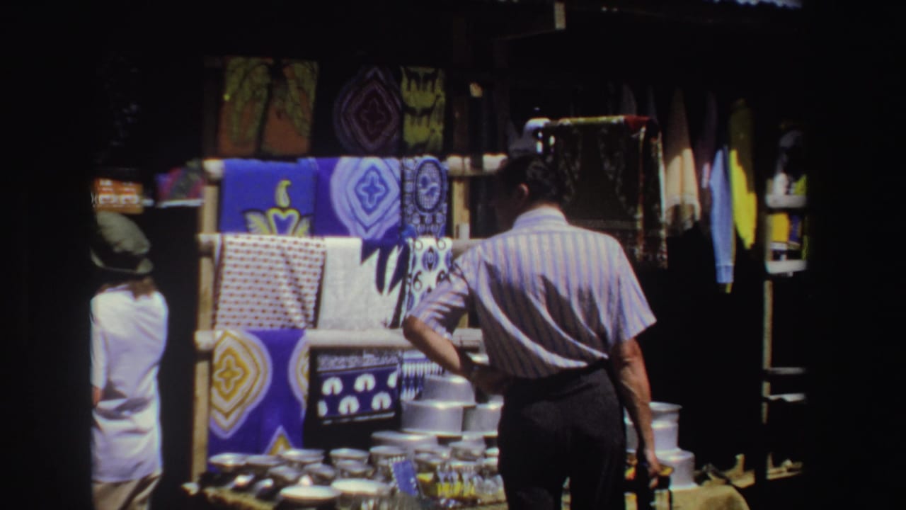 Adults browse a shaded outdoor market stall with colorful textiles and metal goods.