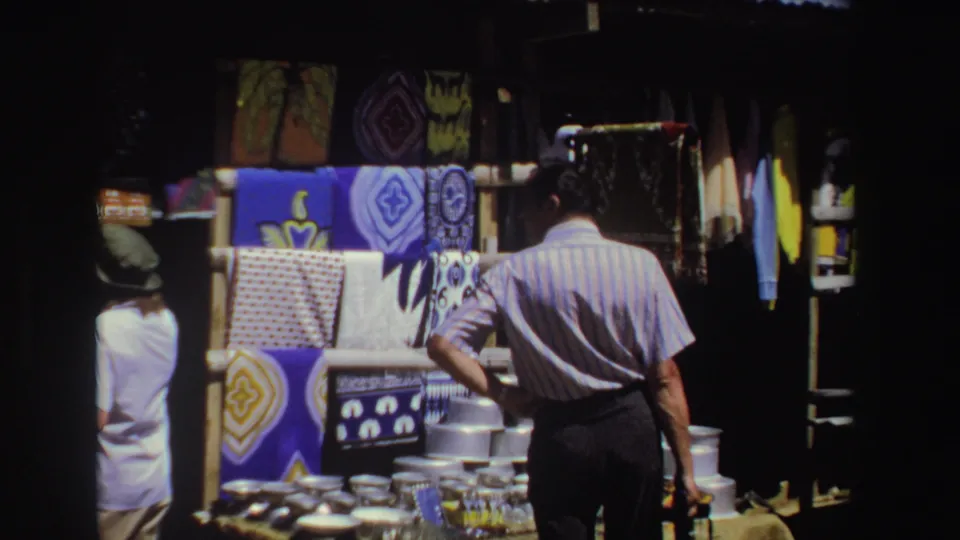 In 1969 South Africa, adults stand near a market stall displaying bright patterned fabrics and stacked metalware.