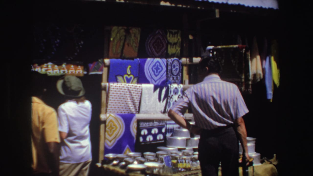 A man and woman stand near colorful fabric panels and stacked metalware at a stall.