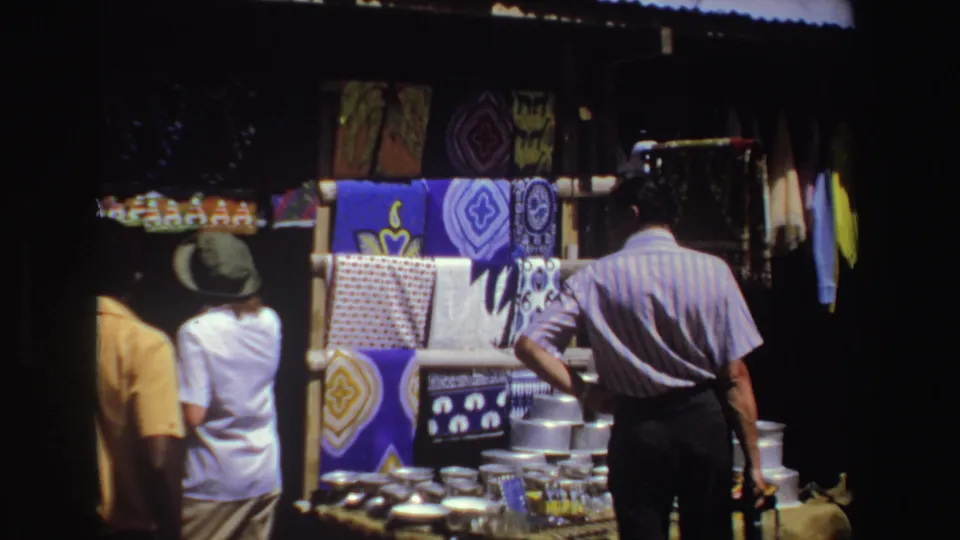 Adults browse a South African outdoor market stall in 1969, with blue and purple textiles hanging behind metal goods.