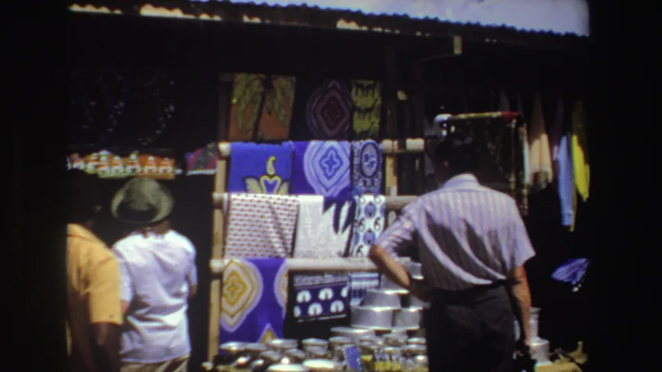 The home-movie frame shows adults browsing a 1969 South Africa market display of patterned fabric and metal cookware.