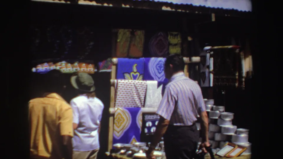 At a 1969 South Africa outdoor market, a man passes a stall with bright textiles, stacked metalware, and displayed small items.