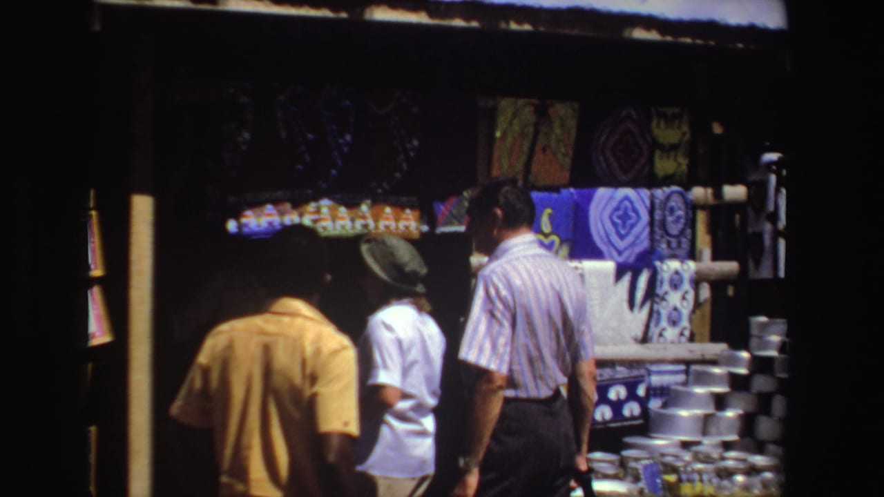 Tourists stand near a shaded market stall with patterned textiles and metal bowls.