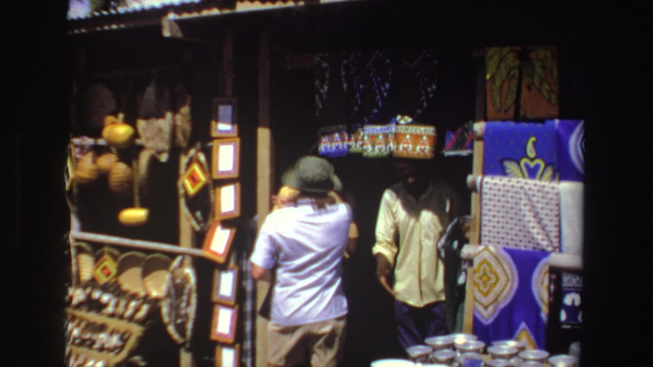 A tourist in a hat faces a vendor beside textiles, baskets, and metal bowls.