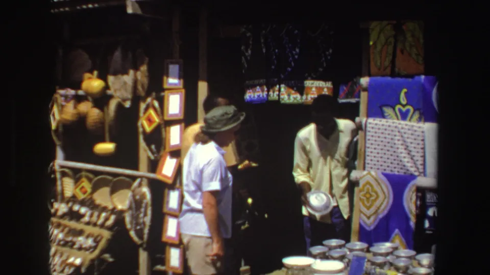 1969 South Africa 8mm frame of a vendor holding a metal bowl as a tourist looks toward a craft and textile display.