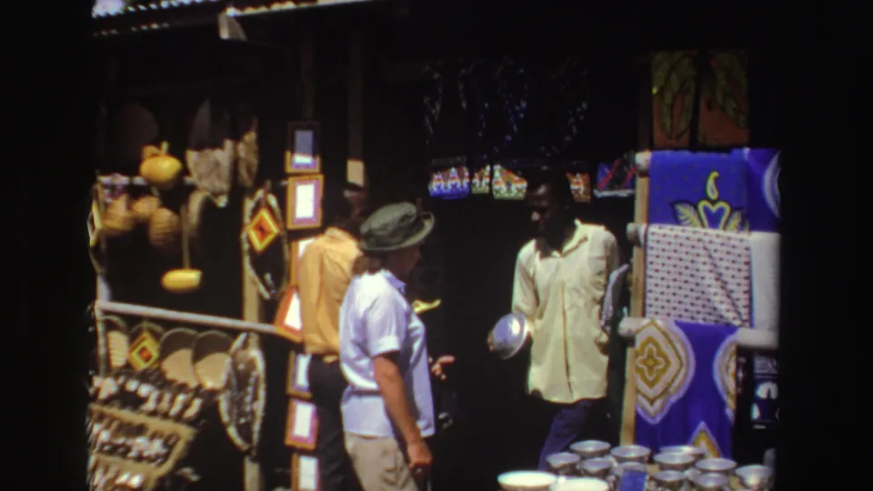 1969 South Africa market frame showing tourists and a vendor amid patterned textiles, woven goods, and metal bowls.