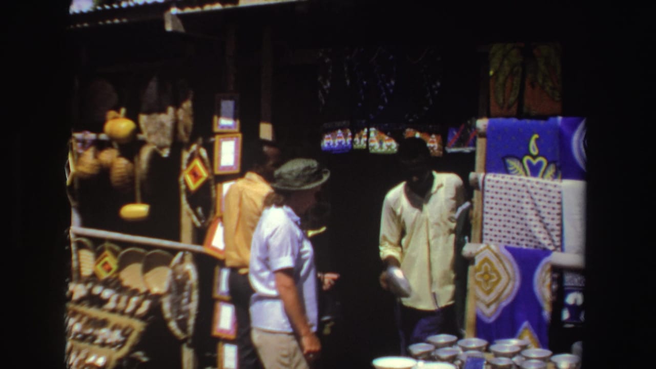 In 1969 South Africa footage, a tourist in a light shirt and bucket hat stands near a merchant at a shaded market stall with bowls and crafts.
