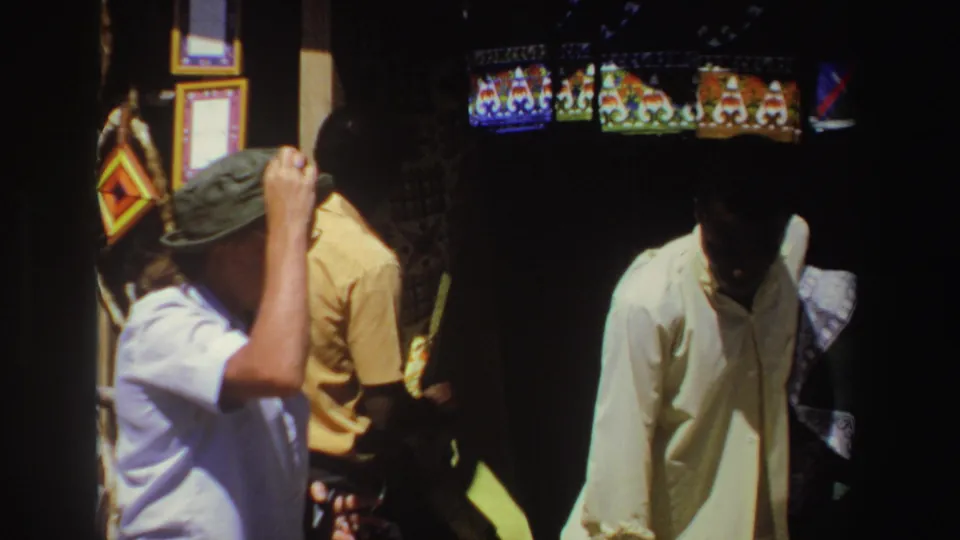 The final frame holds on the tourist and merchant near hanging crafts and bowls, with strong shadow and warm 8mm color from the market setting.