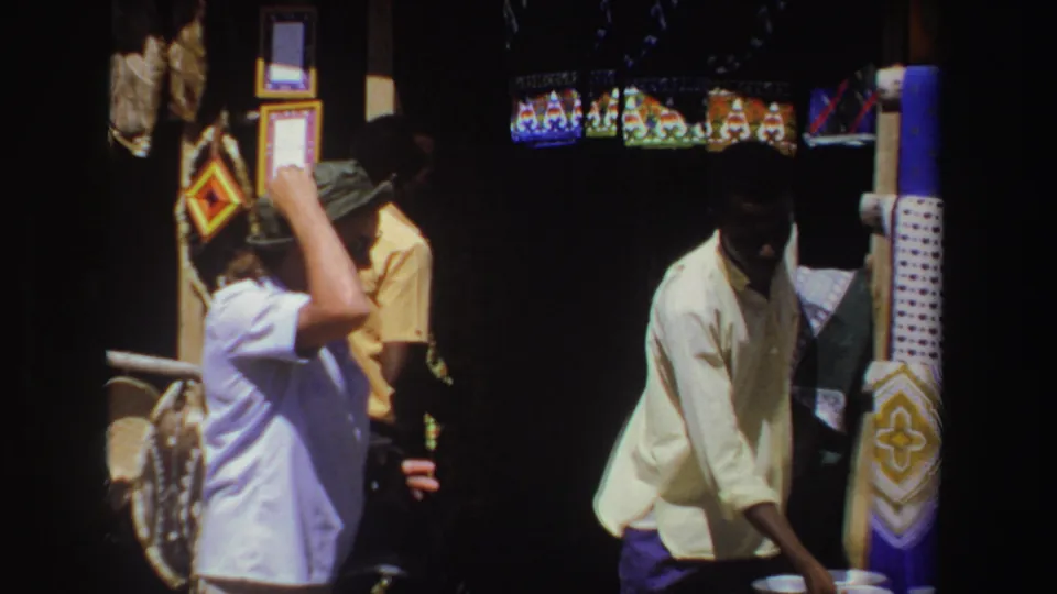1969 South Africa market frame with a young vendor in a light shirt beside patterned cloths and small bowls at a shaded stall.
