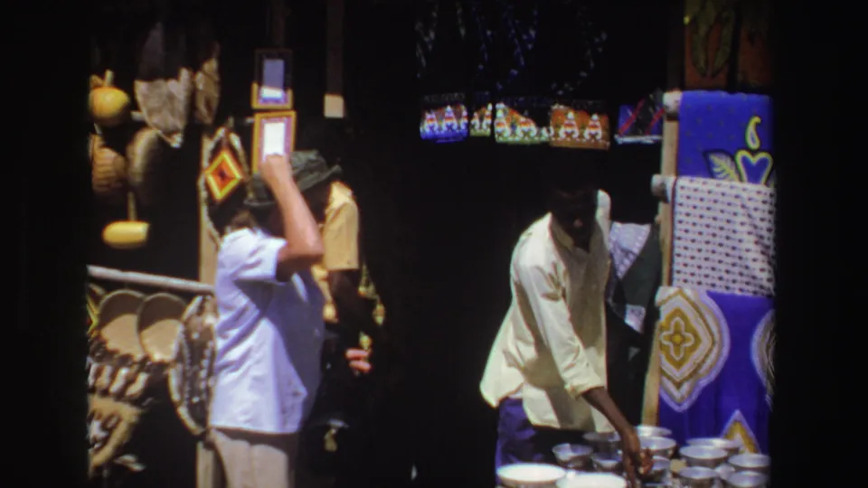 1969 South Africa 8mm frame showing a woman at a craft stall as a young vendor reaches toward a group of small bowls.