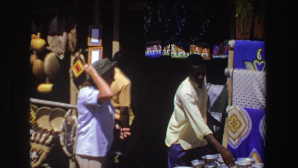1969 South Africa market frame with a young vendor handling bowls beside bright patterned textiles while a woman browses nearby.