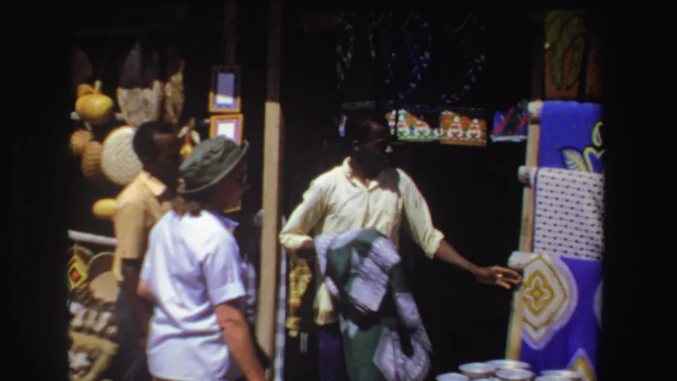 1969 South Africa home-movie still of a merchant gesturing near colorful textiles while a visitor stands by the craft display.
