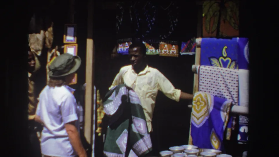 1969 South Africa frame showing a merchant opening or holding a patterned textile beside a visitor at an outdoor bazaar stall.