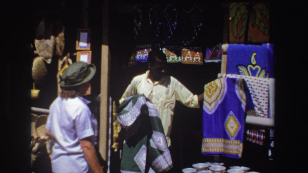 In 1969 South Africa travel footage, a vendor shows blue patterned cloth to a tourist in a green hat beside woven goods.