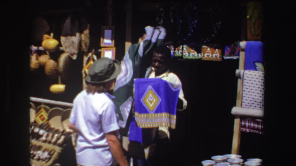 The vendor raises fabric near a blue patterned textile as the tourist stands in front of woven market goods.