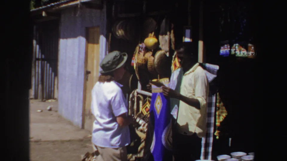 A tourist in a green hat faces a vendor holding blue cloth beside woven goods in the South Africa market scene.