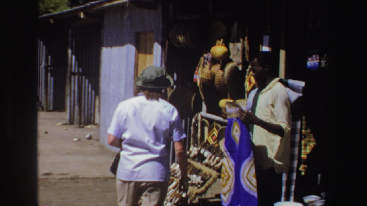 Tourist in a pale blue shirt stands near a craft stall with a vendor and woven goods.