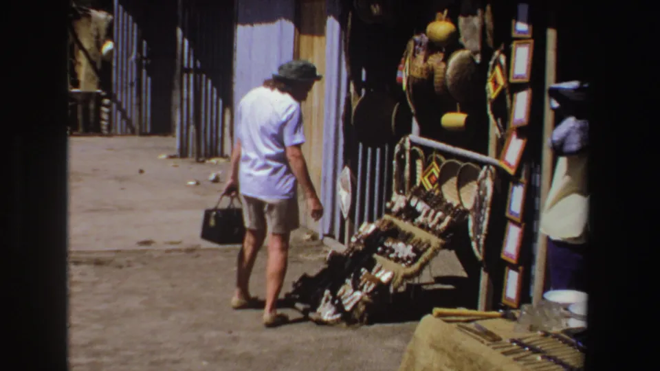 1969 South Africa home-movie frame showing a tourist passing a street-market craft display under strong sunlight.