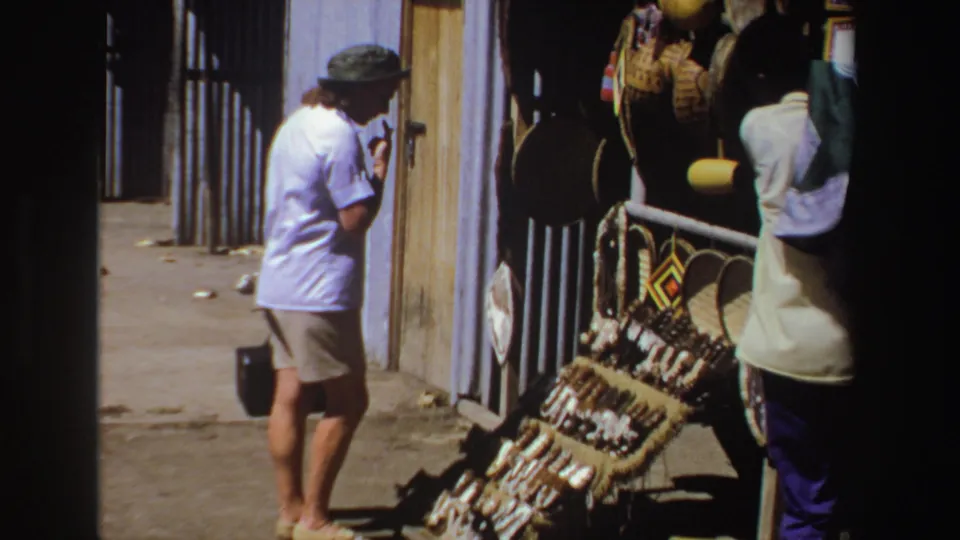 1969 South Africa frame with a tourist near a corrugated building and craft display while a vendor remains partly visible.