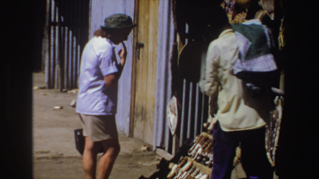 Tourist in a green hat stands near an outdoor craft stall beside a vendor holding woven goods.