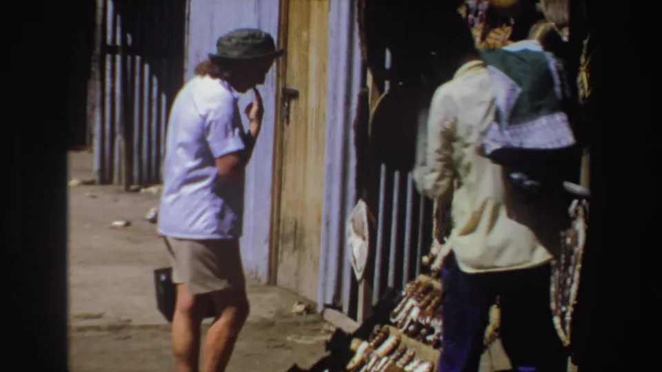 In 1969 South Africa home-movie footage, a tourist in a green hat approaches a sunny outdoor stall where a vendor displays woven crafts.