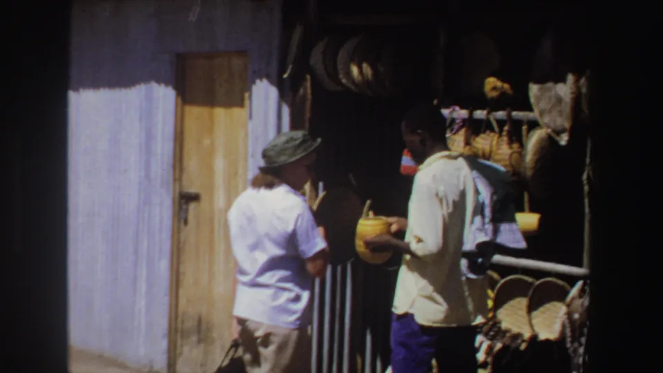 A tourist in light clothing speaks with a vendor holding a yellow woven basket at an outdoor market stall in South Africa, 1969.