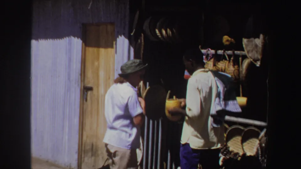 The tourist and vendor continue their exchange beside a wooden door, hanging goods, and stacked woven baskets in the 1969 South Africa footage.