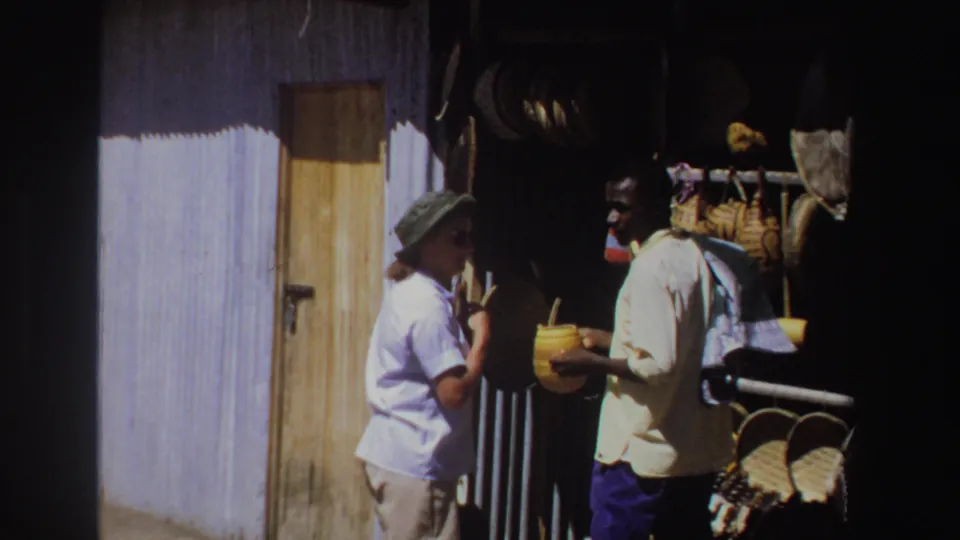 A vendor with a yellow basket faces a tourist in a green hat beside woven crafts and a sunlit wall in South Africa, 1969.