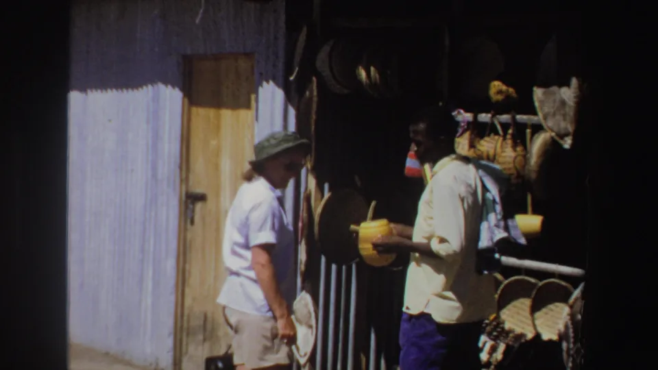 1969 South Africa 8mm frame of a woman in a brimmed hat facing a vendor with woven goods hanging at a market stall.