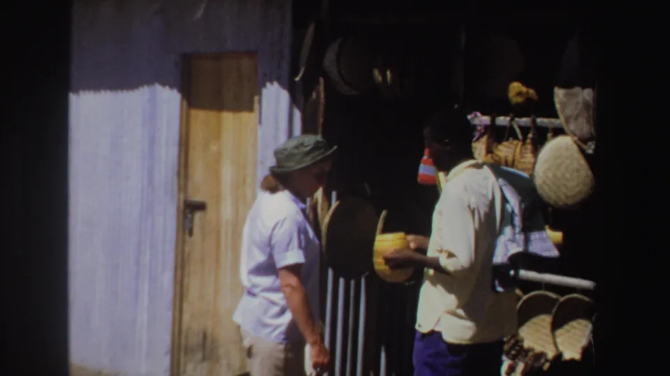 1969 South Africa market view with a woman considering an item held by a vendor beside hanging baskets and a wooden door.