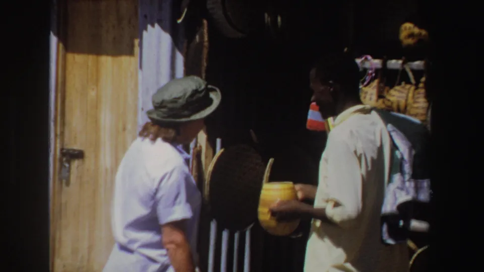 Close 8mm frame from 1969 South Africa showing a market exchange between a woman in a hat and a vendor holding a craft item.