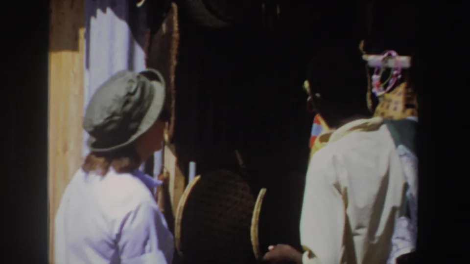 1969 South Africa home-movie frame with the market figures partly turned from camera beside shadowed craft displays.